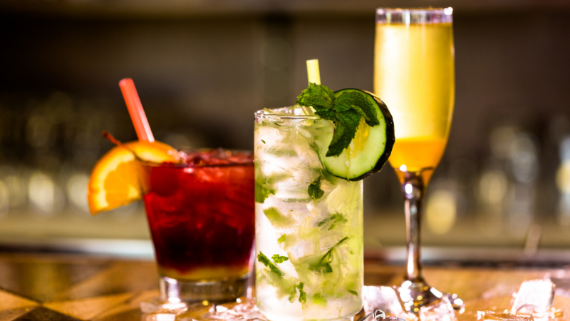 Three different cocktails on a bar counter. A red drink in a rocks glass, a green herby drink in a highball glass and an orange drink in a flute.