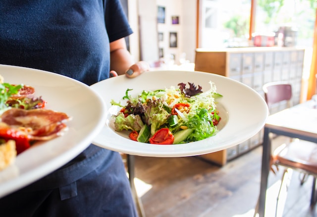 Person in apron holding two plates of salad with table in background
