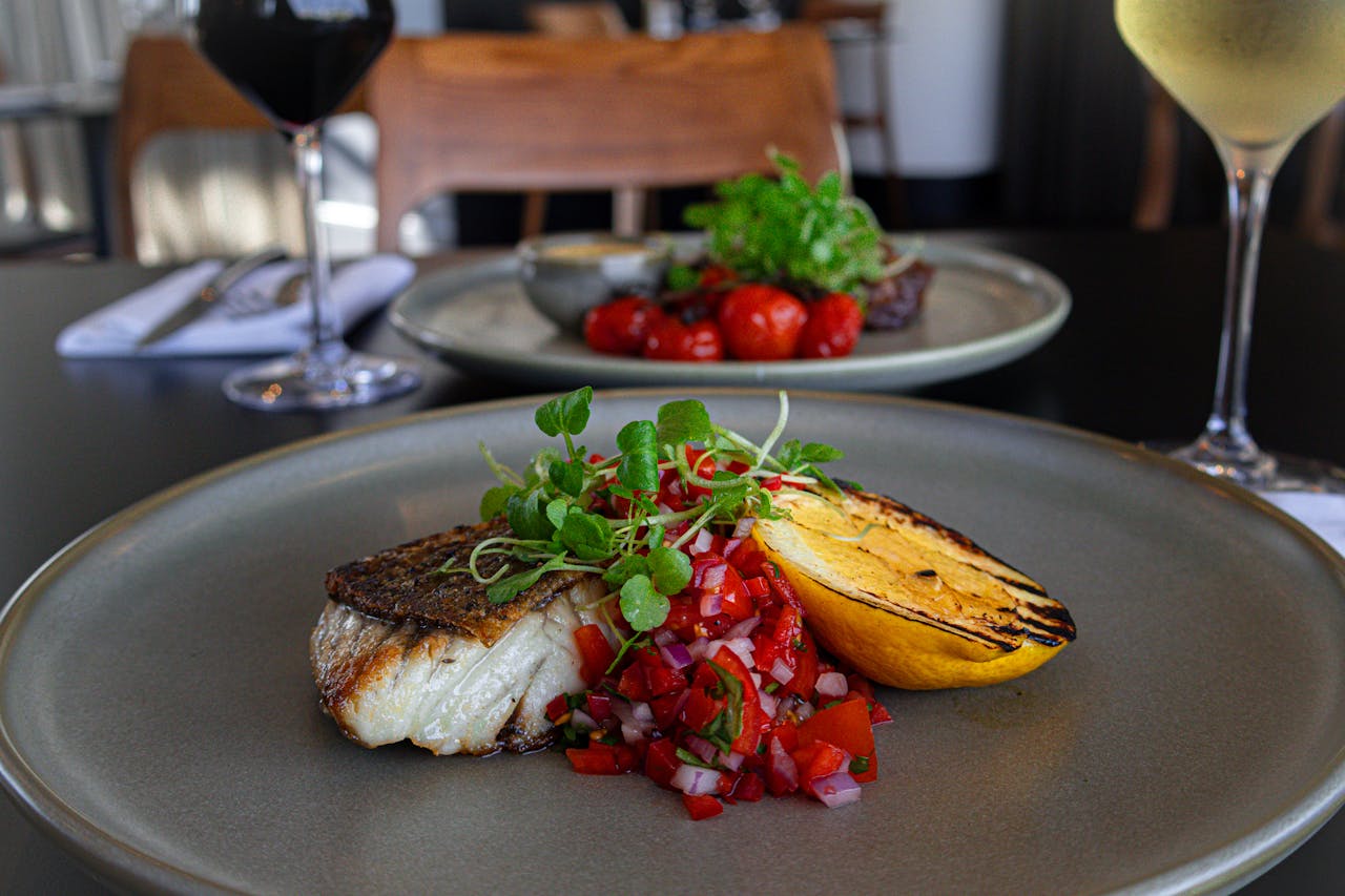 Fish, salsa and grilled lemon on plate in restaurant with glasses of wine