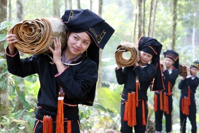 Women in black and red garments carrying rolled up canvas on their shoulders through the forest