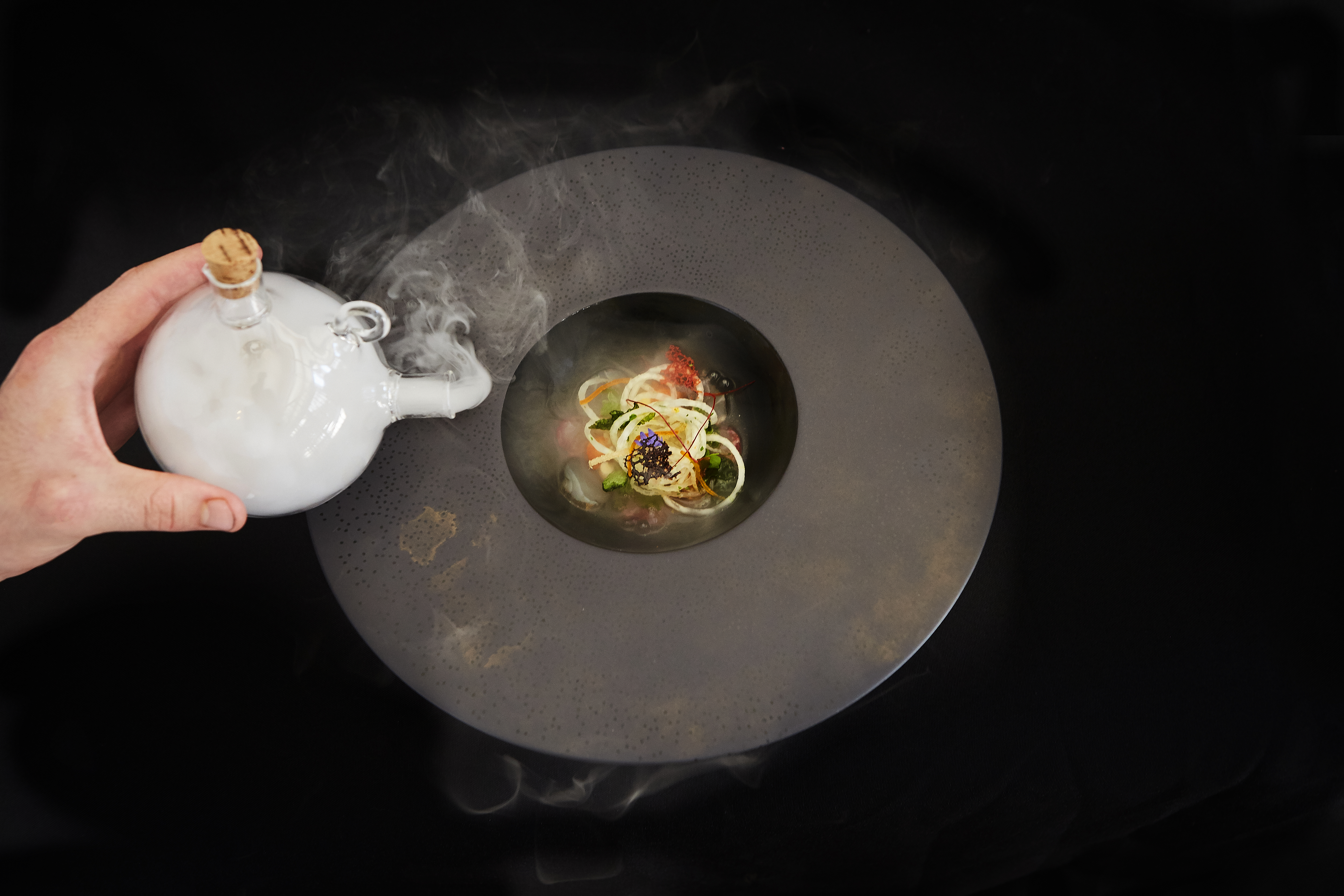 Callan Austin's signature dish. Food in dark bowl against black background. A hand on the left waves smoke over the dish from a stoppered jug.