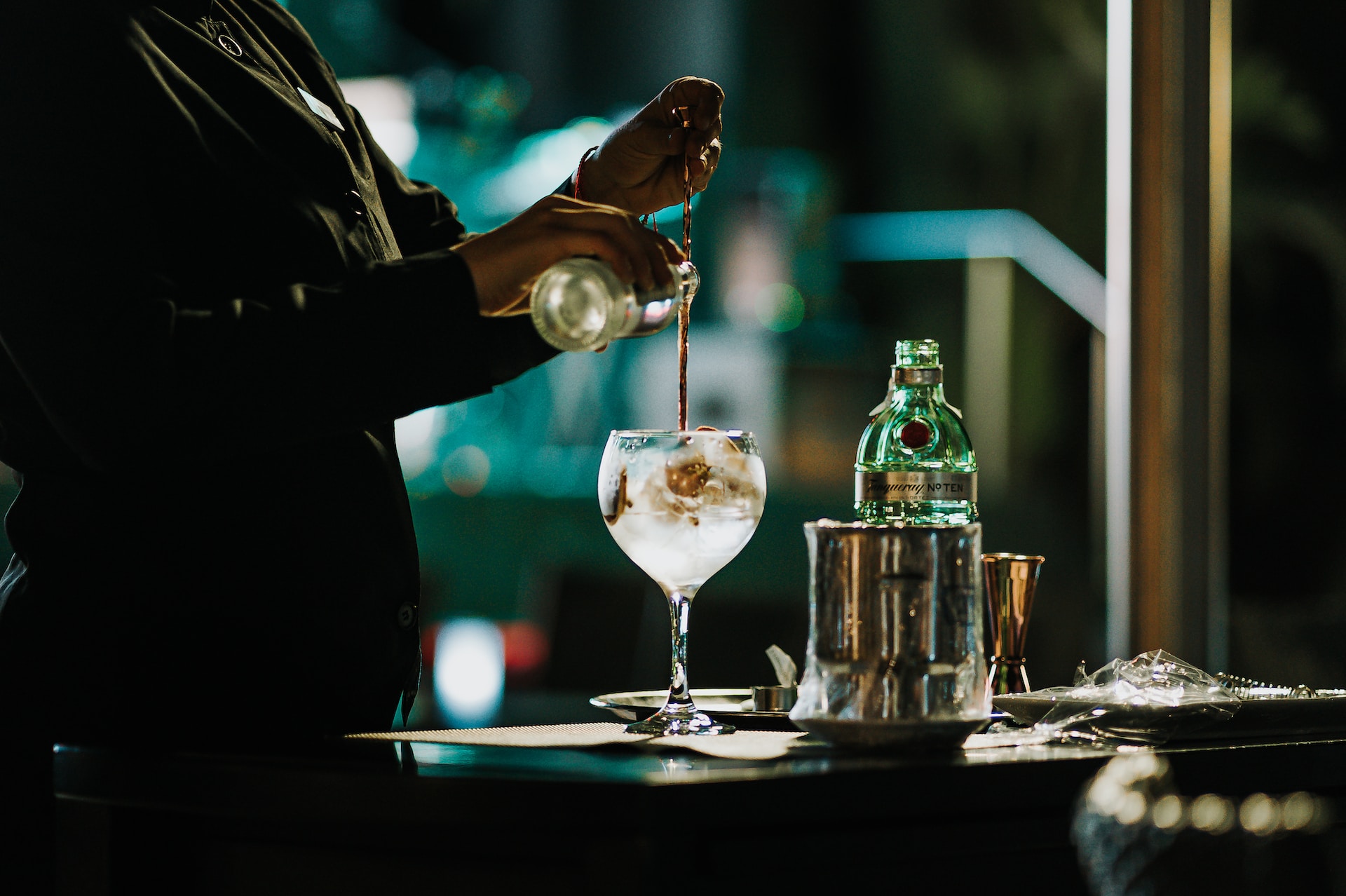 Bartender making gin cocktail in dimly-lit bar, stirring liquid in balloon glass and pouring tonic in