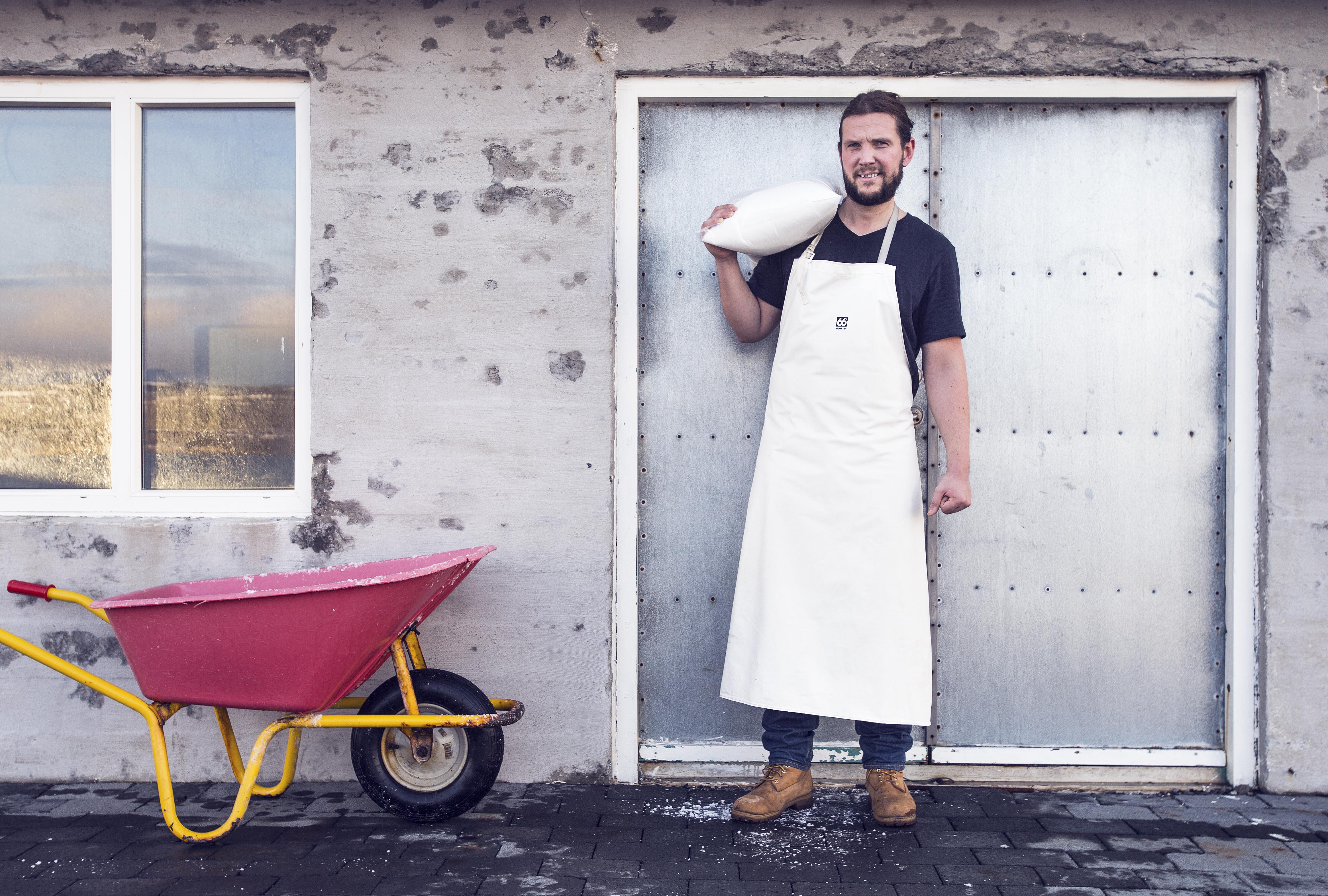 Björn Steinar Jónsson stands outside door of Saltverk, wearing an apron, carrying a bag of salt on one shoulder. A red wheelbarrow is beside him.