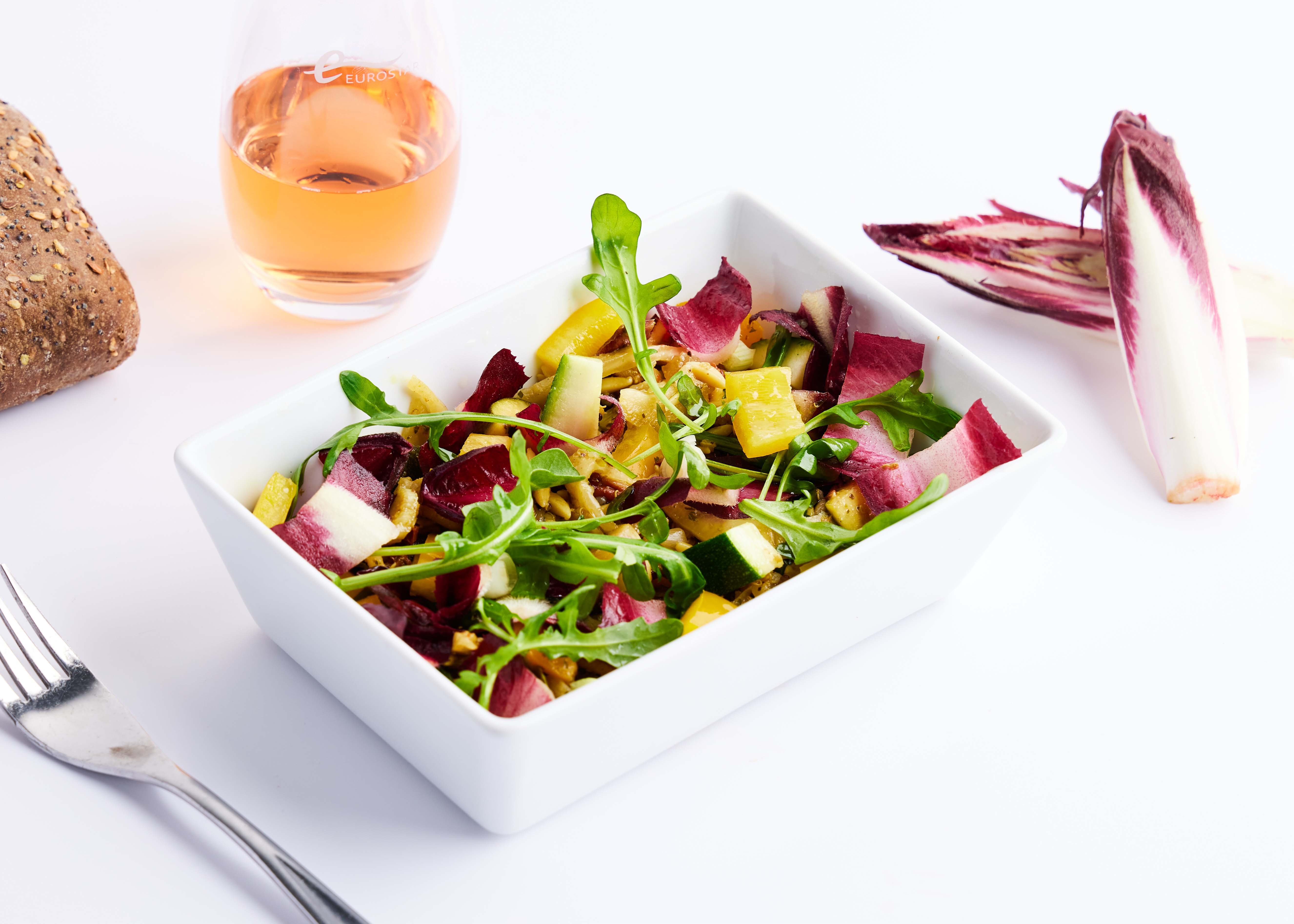 Small square bowl of salad on white surface with glass of orange liquid, fork, bread and radicchio leaves