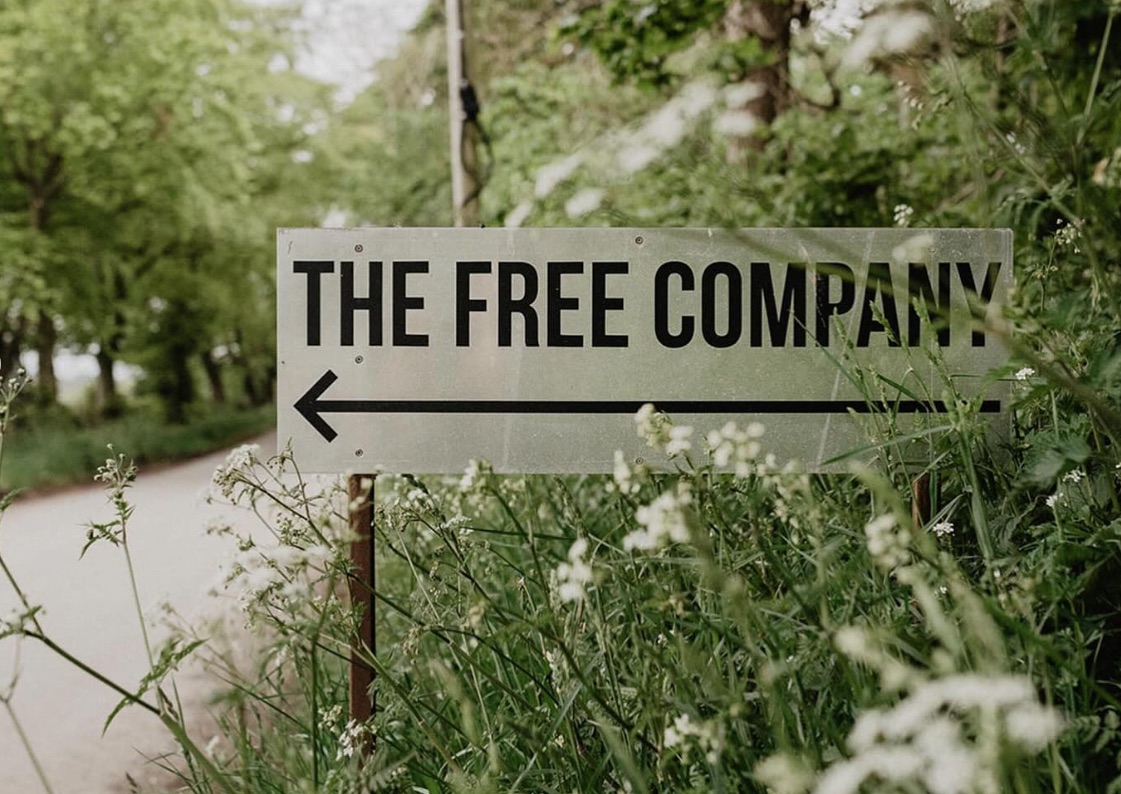 A road sign in a green hedgerow verge saying 'The Free Company' with an arrow pointing left