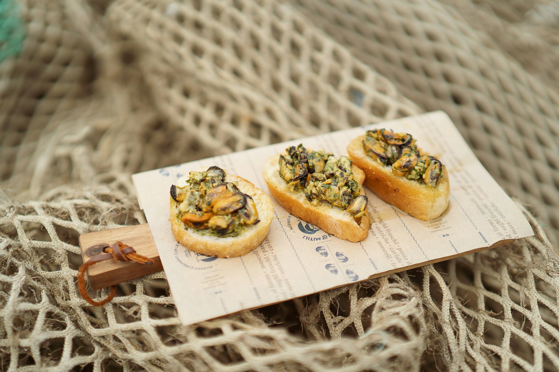 Mussels on sliced baguette placed on a paper-covered board, shot on top of a beige net