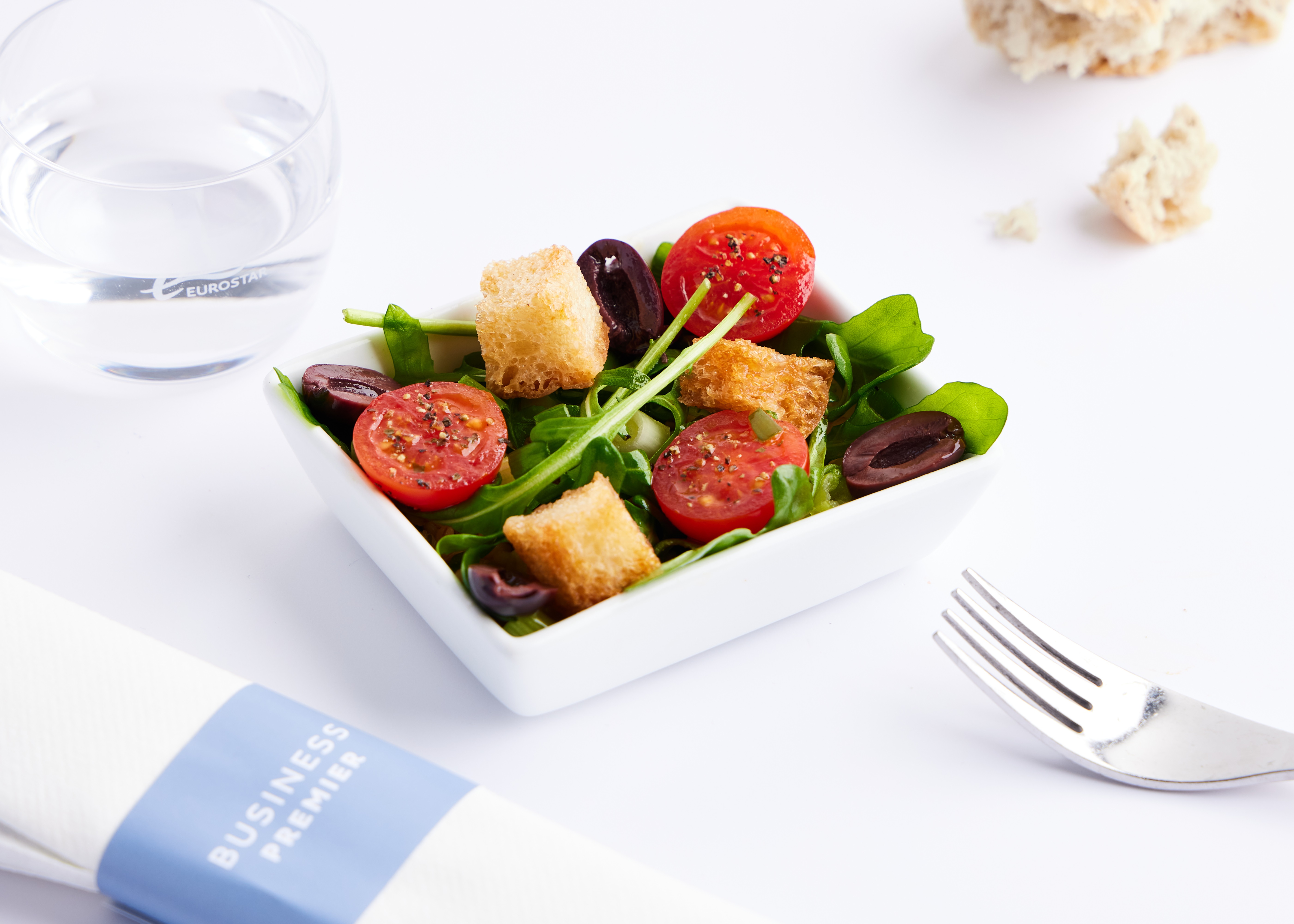 Small square bowl of salad on white surface with napkin and fork
