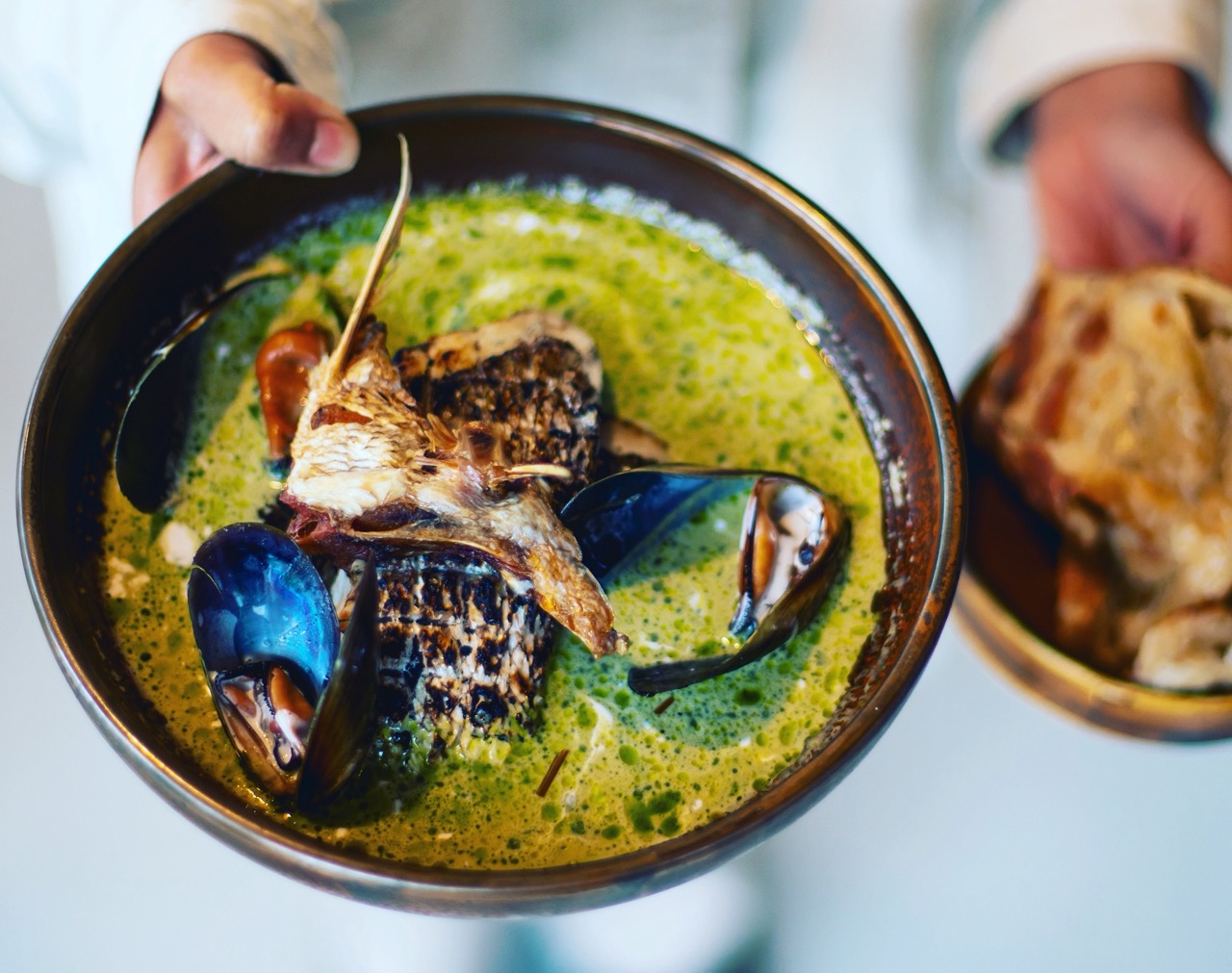 Hands holding two bowls, one with green curry, one with bread